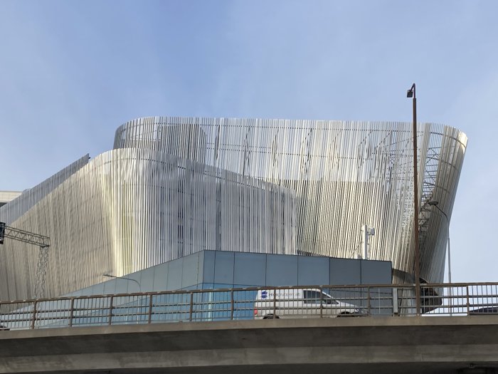 Modern byggnad med ovanlig arkitektur och metallfasad under en klar himmel, nära en bro eller viadukt.