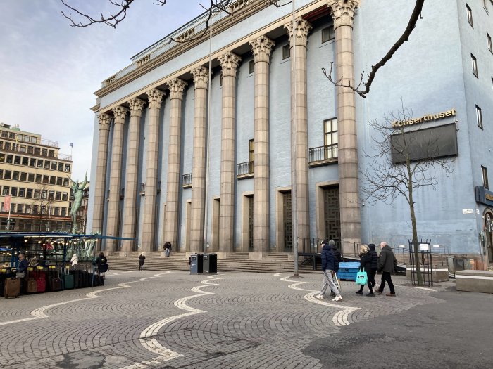 Storslagen byggnad med kolonner, människor på torg, stadsmiljö, molnig himmel, "Konserthuset" skylt.