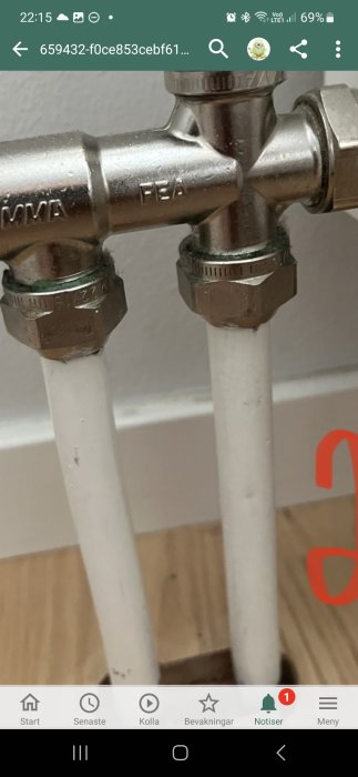 Närbild på kromad T-rördel med anslutna vita vattenrör mot en vägg.