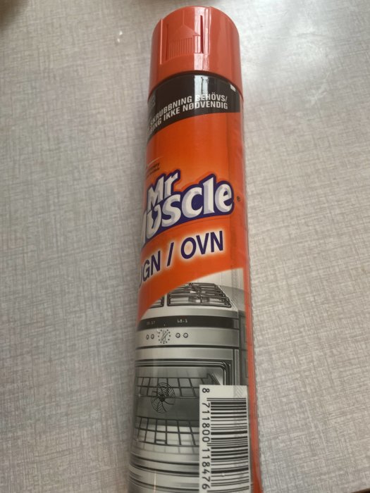 Flaska med Mr Muscle rengöringsmedel för ugn, stående mot en texturerad yta.