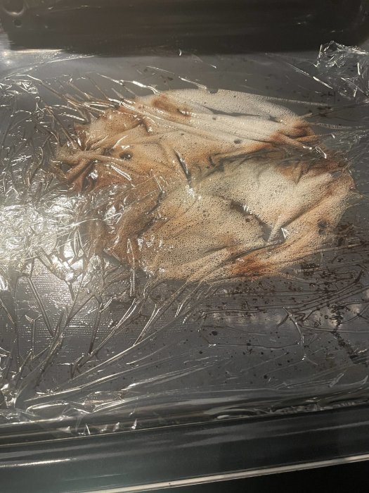 Ugnsform med fisk under plastfolie, tillagning i process, ångbildning, synlig vätska, köksmiljö.