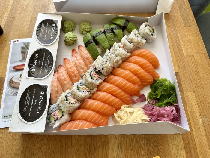 En öppen sushi take-away låda med nigiri, maki, sashimi, sojasås, ingefära, och wasabi.