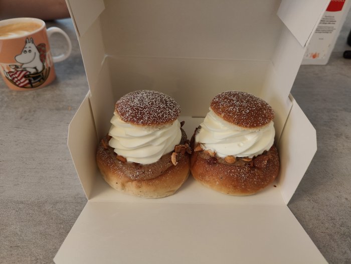 Två semlor med grädde och mandelmassa i en pappersförpackning, nära en kaffemugg.