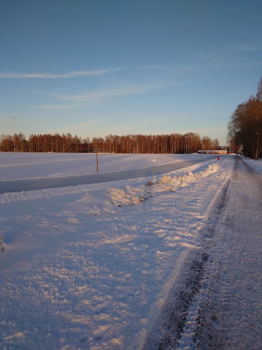 Snötäckt landskap vid solnedgång med fotspår och väg. Träd i bakgrunden, klar himmel.