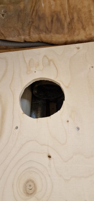 Utskuret hål i träplywood, avslöjar del av mörk ventilationsslang, praktisk konstruktion, grov finish.