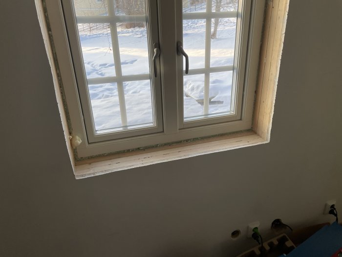 Ett fönster med utsikt över snötäckt landskap, inomhus, ofärdig fönsterkarm, solljus, grå vägg.