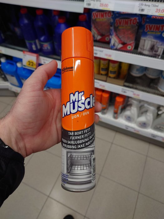 Hand håller en Mr Muscle rengöringsspray i en butik, hyllor med rengöringsmedel i bakgrunden.