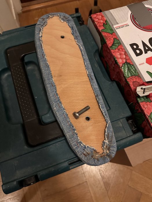 Skateboard utan hjul, med jeans-tygkant, ligger på en grön verktygslåda bredvid en låda.