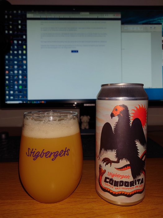 Ett skumfyllt glas öl framför en burk med etikett "Condordita", bakom en datormonitor med text.
