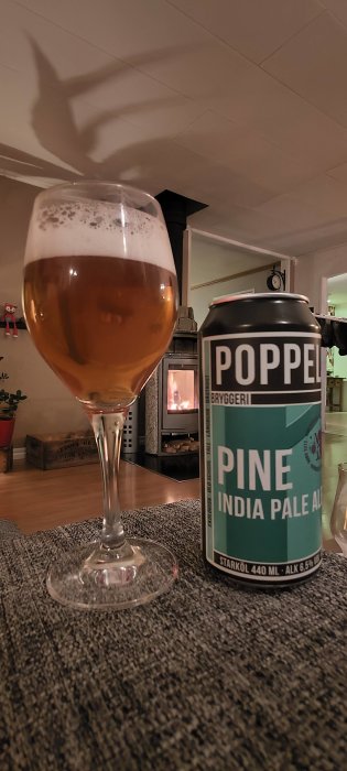 Ölglas med skumkrona, burk India Pale Ale, mysig brasa i bakgrunden, hemtrevlig atmosfär.