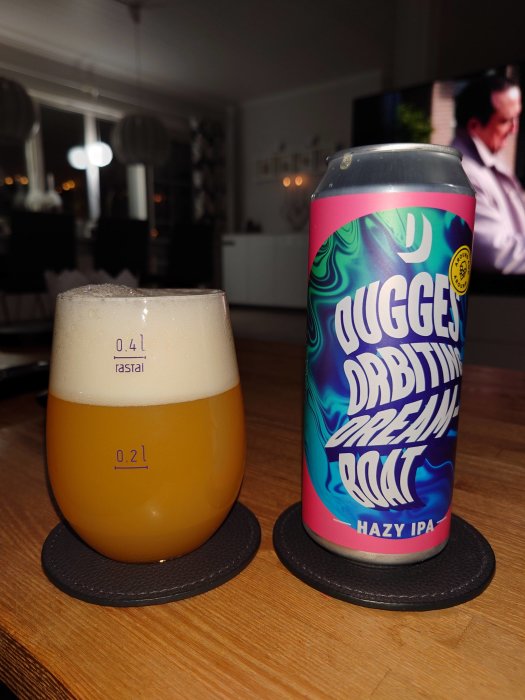 Ölglas fyllt med skum och hazy IPA, burk med färgstark etikett, inomhusmiljö, tv-skärm i bakgrunden.