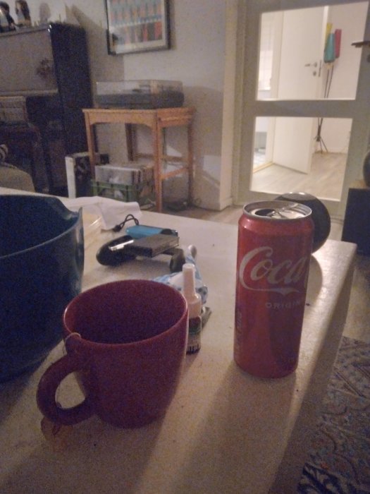 Inomhusmiljö, bord med mugg, Coca-Cola-burk, fjärrkontroll, rumsinredning, vardagsrum, dämpad belysning, kvällstid.