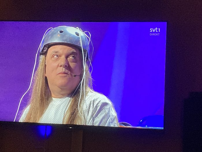 Person med hjälm, mikrofon, förundrad blick. TV-skärm, SVT1 Direkt-logotyp, lila bakgrund.