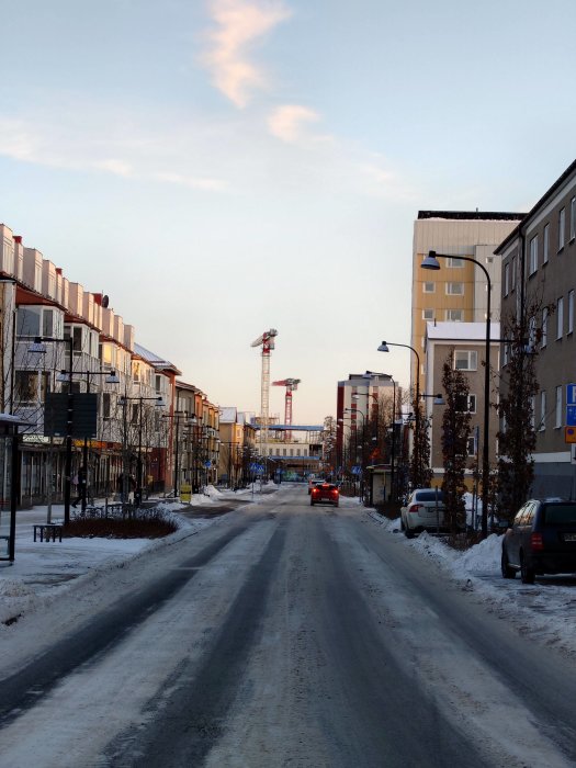 Snötäckt gata, bostadshus, blå himmel, kran i fjärran, bil, gatlyktor, vinterdag.