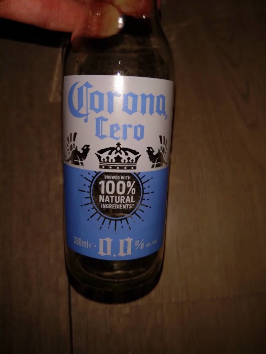 En hand håller en Corona Cero-flaska, alkoholfritt öl, med texten "100% NATURAL INGREDIENTS", mörk bakgrund.
