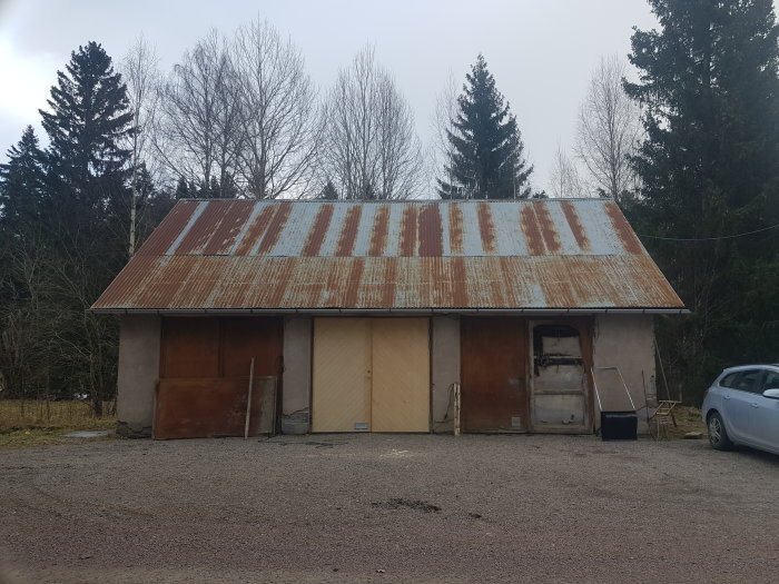 Äldre lada, rostig plåttak, två garageportar, en dörr, träd, grå himmel, parkerad bil.