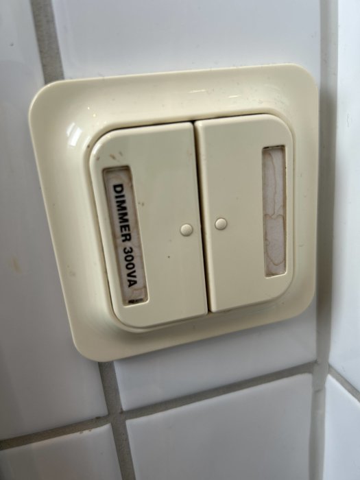 Beige väggströmbrytare med dimmerfunktion, smutsig, monterad på vit kakelvägg.