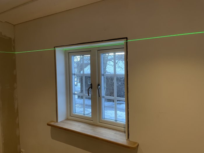 Ett fönster, snöig utsikt, grönt laserverktyg visar linje för nivellering under renovering.