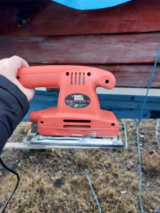 En hand håller en orange Black & Decker slipmaskin utomhus nära en trävägg.