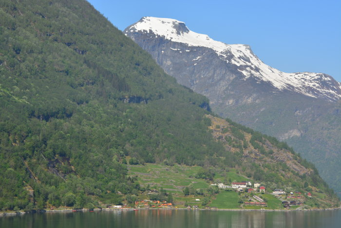 Norsk fjord, snötäckta bergstoppar, gröna träd, spegelblankt vatten, pittoresk by.