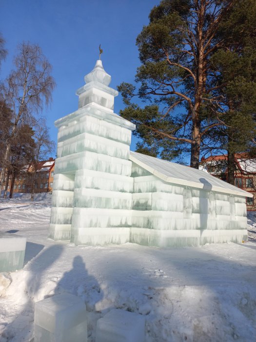 Struktur byggd av isblock mot en klarblå himmel, snöigt landskap, träd i bakgrunden. Skugga av en person syns.