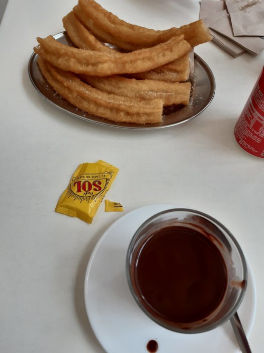 Tallrik med churros, kopp med varm choklad, sockerpacket, Coca-Cola burk, vita ytor.