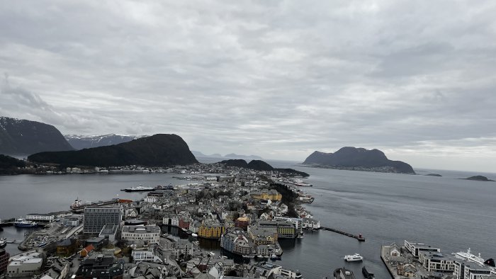 Stad vid kusten, molnig himmel, omgiven av berg och vatten, färgglada byggnader, nordiskt landskap.