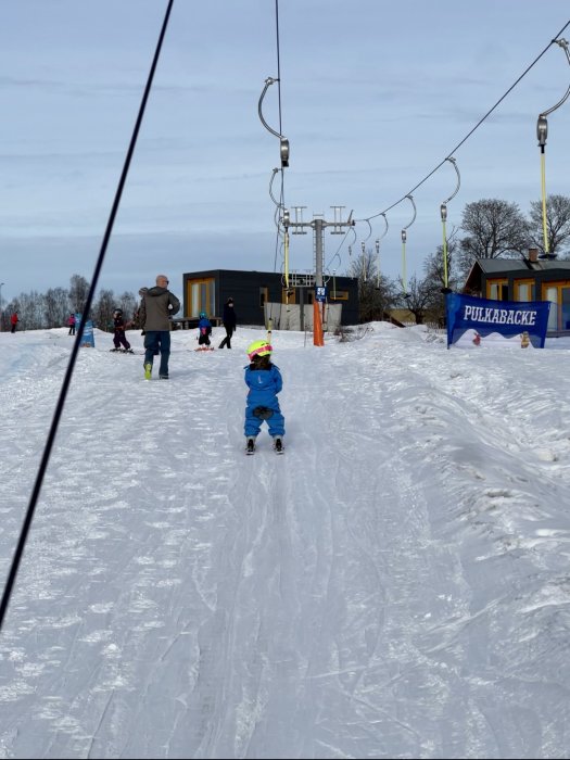 Ett barn åker skidor i en släplift, människor i en snöig backe, "Pulkabacke" skylt.