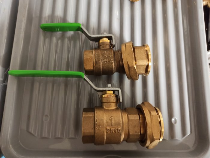 Två mässingskulventiler med gröna spakhandtag på grå plastbakgrund. VVS-utrustning eller installationstillbehör.