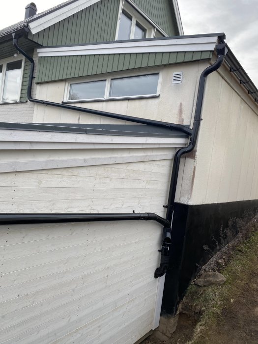 Vit husfasad med svarta stuprör, fönster, takränna, vädringslucka, delvis synlig grund.