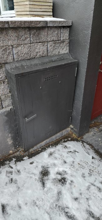 Svart metallucka i vägg, snötäckt mark, ventilationshål, utomhusmiljö, kallt väder.