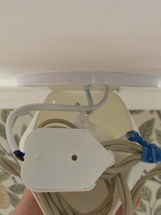 Taklampa under installation, vit takkåpa, grå textilkabel, någon håller i lampan.