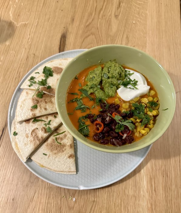 Mexikansk soppa med avokado, gräddfil, majs, bönor, chili; serverad med quesadillas. Färgglatt, aptitretande, på ett träbord.