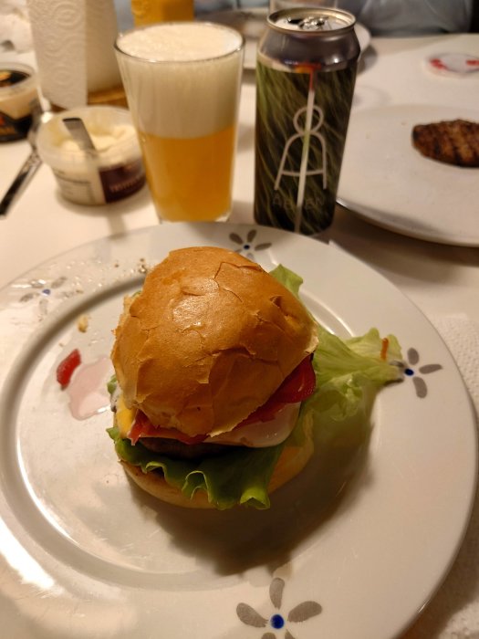 Hamburgare med sallad och tomat på tallrik, ölglas, burk, bakgrundsgrilltillbehör. Hemmamiljö.