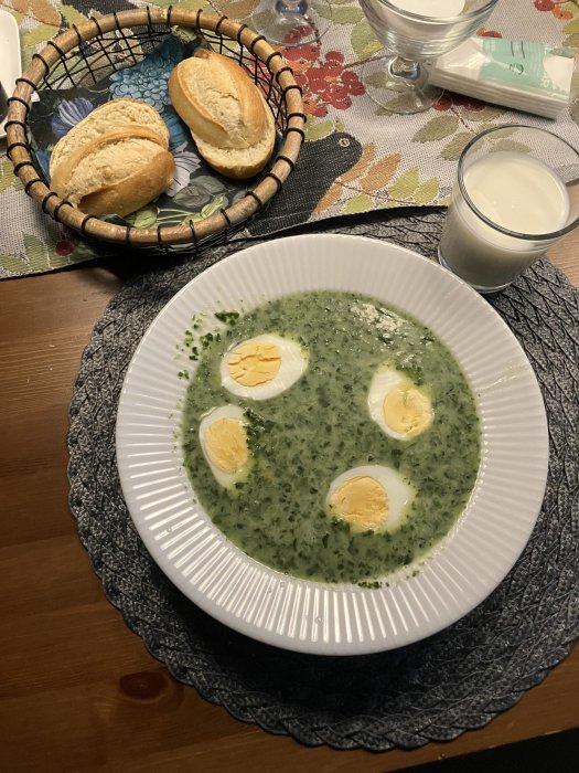 Tallrik med grön ärtsoppa och skivade ägg, korg med bröd, mjölkglas, mönstrad duk på träbord.