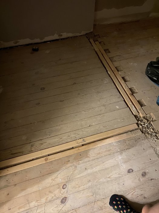 Renoveringsarbete, upprivna golvlistor, trägolv, synliga spikar, någons fot i hörnet, byggavfall.