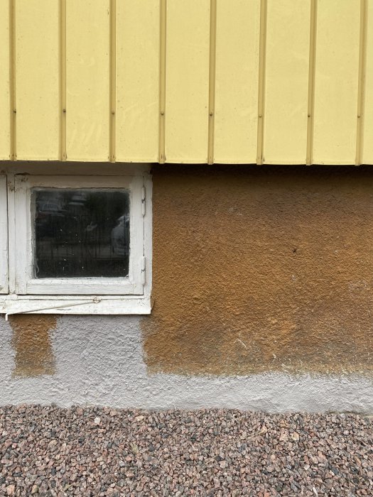 Gulfärgad vägg, vitt fönster, brunaktig puts, fuktfläckar, grusgrund. Kontraster i textur och färg. Exteriör arkitektur närbild.