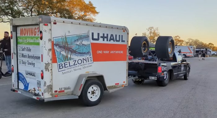 En U-Haul flyttbil och en pickup med stora däck på flaket, människor i bakgrunden, klar himmel.