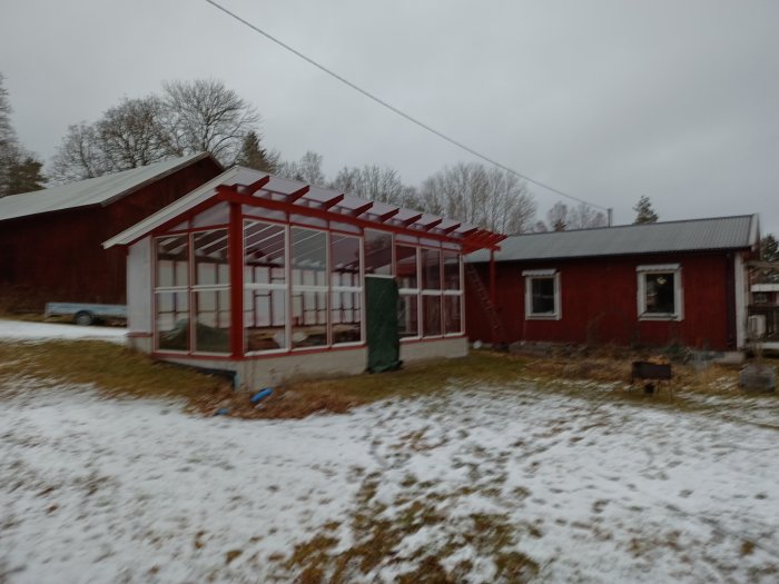 Ett rött hus med glasväxthus, snötäckt mark, grå himmel och lantlig miljö.