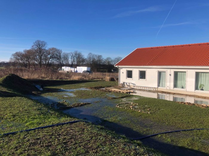 Nybyggd villa med röda tegeltak, översvämmad trädgård, landsbygd, klart himmel, pågående markarbeten, stillastående vatten.