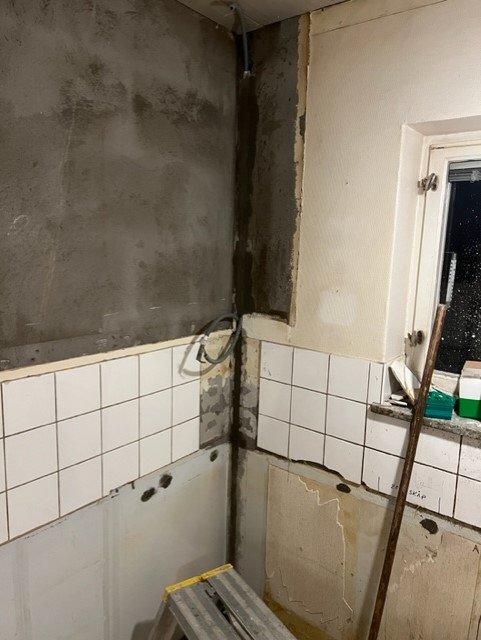 Ett badrum under renovering med avlägsnade kakelplattor, utsatta rör, och ojämnt putsade väggar.