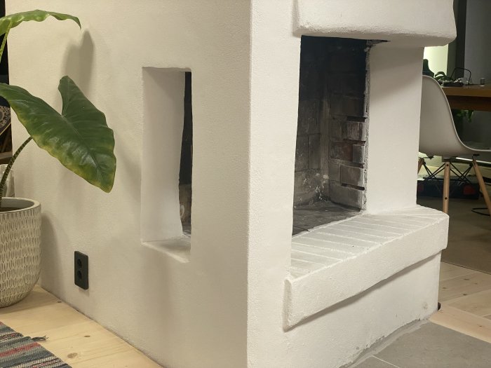 En renoverad öppen spis med vita putsade väggar, ett tomt eldstadsutrymme och en planta till vänster.