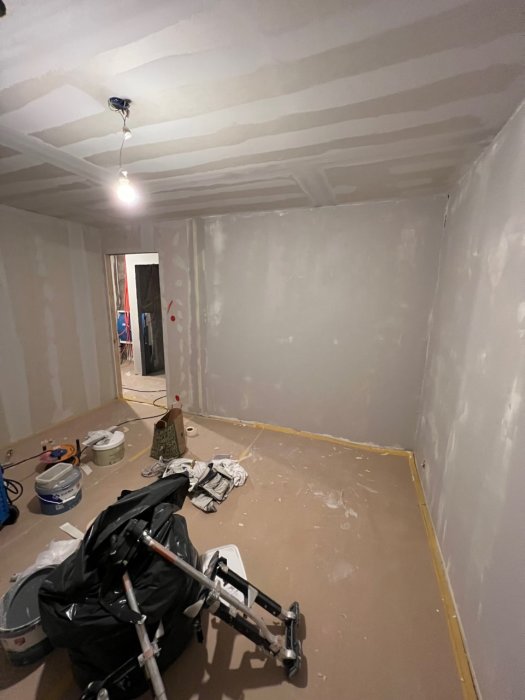 Ett rum under renovering med oslipade gipsväggar, målarförråd och en ihopfälld barnvagn på golvet.