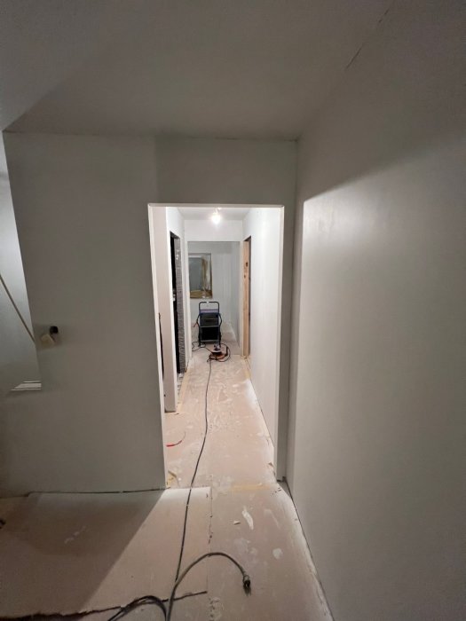 Lång, smal korridor i renovering, kabeldragning på golv, vita väggar, avskalat, arbetsbelysning längre in.
