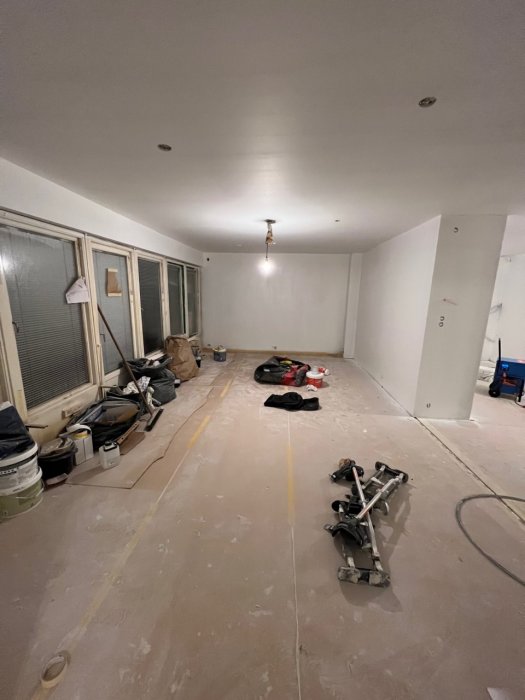 Ett rum under renovering med byggmaterial, skyddspapp på golvet, och vitmålade väggar.
