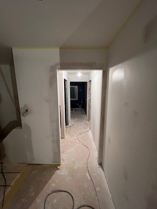 En korridor under renovering med oslipade väggar och golv, öppna dörrkarmar samt kablar på golvet.