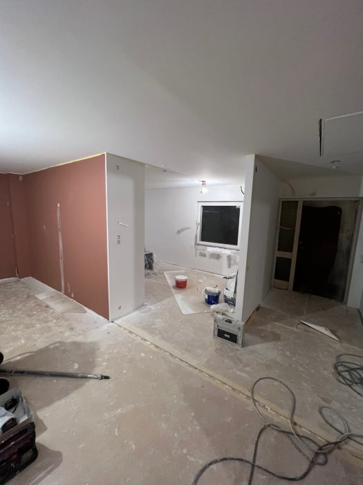 Renoveringsarbete pågår inomhus, ommålad vägg, oskyddad golv, byggmaterial och verktyg synliga.
