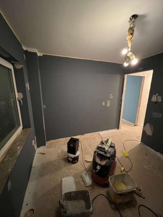 Ett rum under renovering med målargrejer, skyddad golv, målad vägg, osatt taklampa och en stängd dörr.