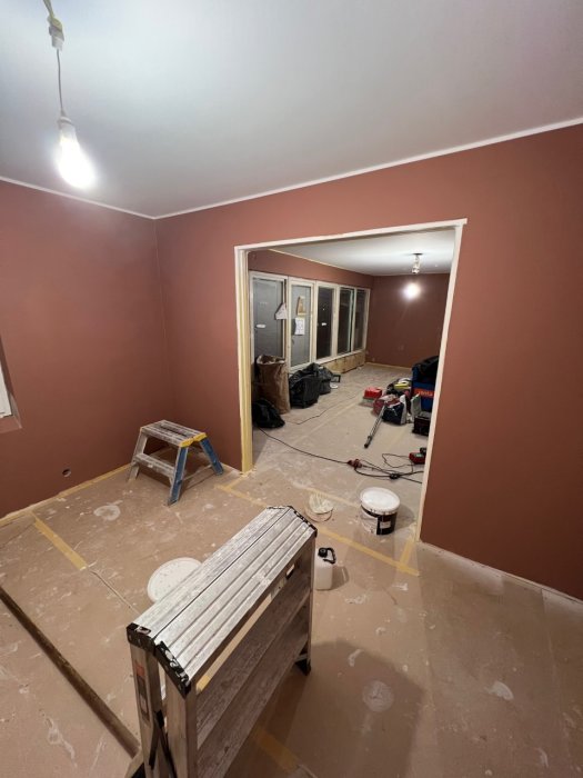 Renovering pågår, rosa väggar, täckt golv, stegar, arbetslampor, dammigt, ombyggnadsprocess, oinredda rum, sladdar, efterarbetskaos.
