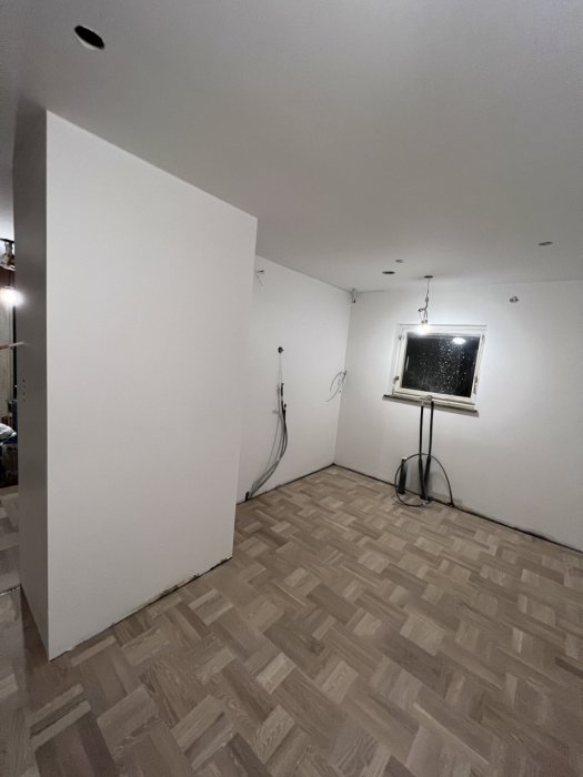 Ett tomt rum under renovering med parkettgolv, vit vägg och elektriska installationer.